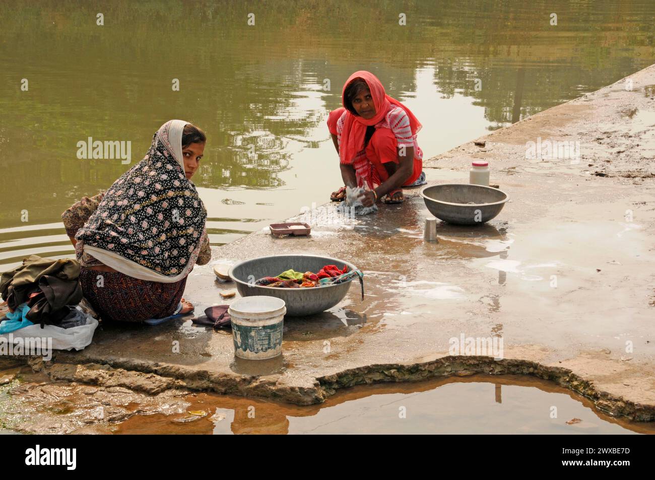 Due donne che lavano il bucato e i piatti sulla riva del fiume in abiti tradizionali, Udaipur, Rajasthan, India del Nord, India Foto Stock