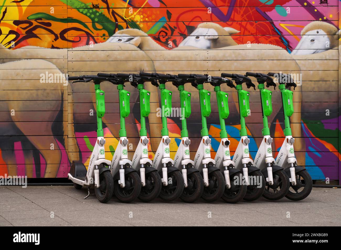 Diversi scooter elettrici parcheggiati nel noleggio bici della società Lime, OnLime e Uber, dietro di esso recinzione di costruzione con pecore, gregge di pecore Foto Stock