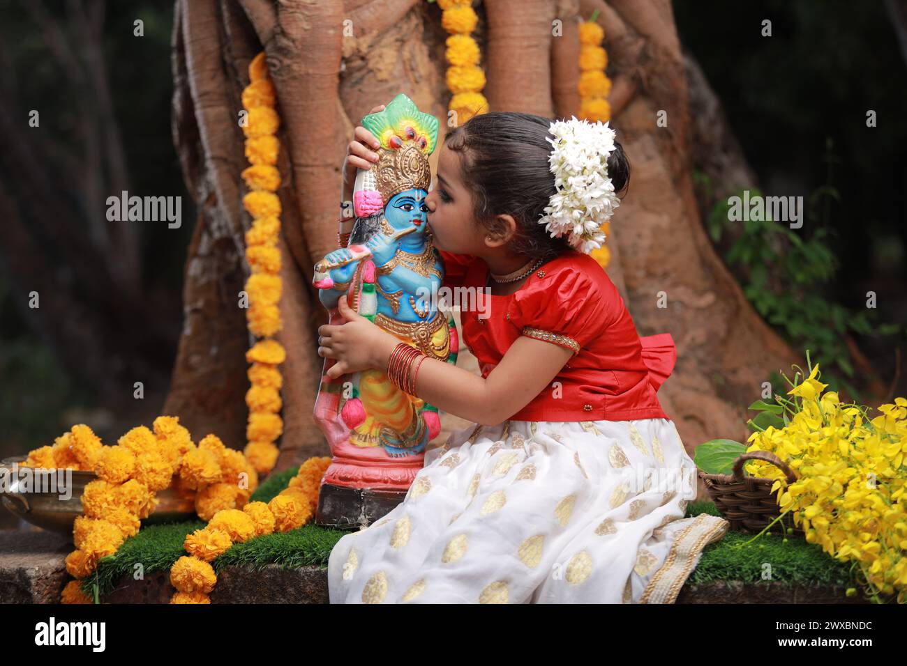 Una bambina carina che indossa un abito Kerala, gonna lunga color oro e camicetta rossa, seduta sotto l'albero banyan con la statua del signore krishna-Golden sh Foto Stock