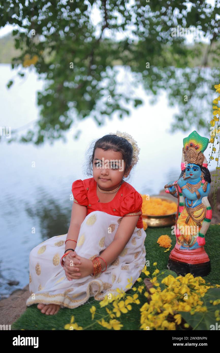 Una bambina carina che indossa un abito Kerala, gonna lunga color oro e camicetta rossa, seduta sotto l'albero banyan con la statua del signore krishna-Golden sh Foto Stock