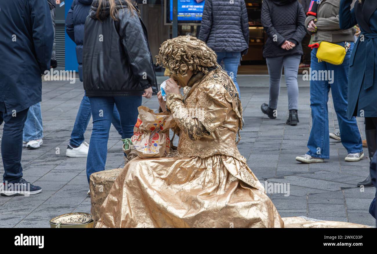 Una statua vivente femminile o umana fa una pausa per mangiare un McDonald's per il pranzo. Un artista di strada fa una pausa pranzo tra gli amanti dello shopping. Foto Stock