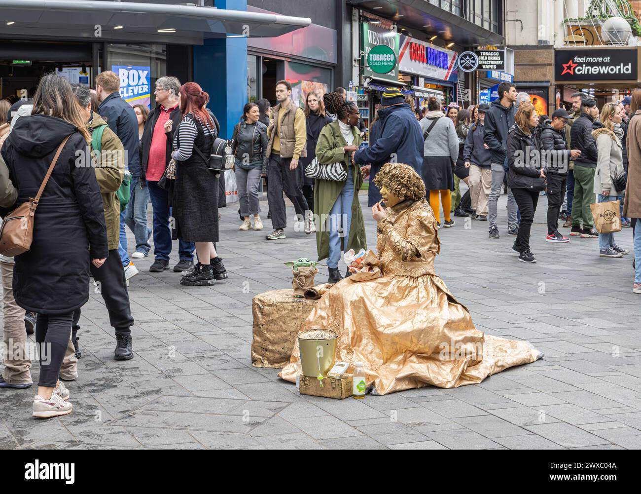 Una statua vivente femminile o umana fa una pausa per pranzare mentre la gente guarda. Un artista di strada guarda il mondo passare mentre pranza Foto Stock