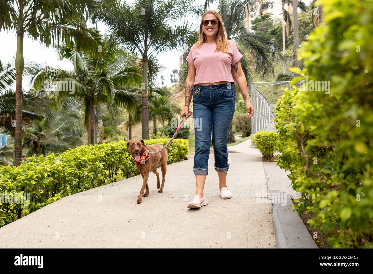 Una donna di mezza età sorride e guarda la telecamera mentre cammina con il suo cane, che conduce al guinzaglio attraverso un parco. Foto Stock