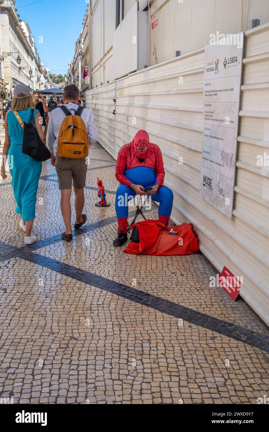 Uomo grasso in sovrappeso vestito da ragno in una strada di Lisbona, seduto a guardare il suo cellulare, vendendo figure di ragni grassi con il passaggio dei turisti Foto Stock