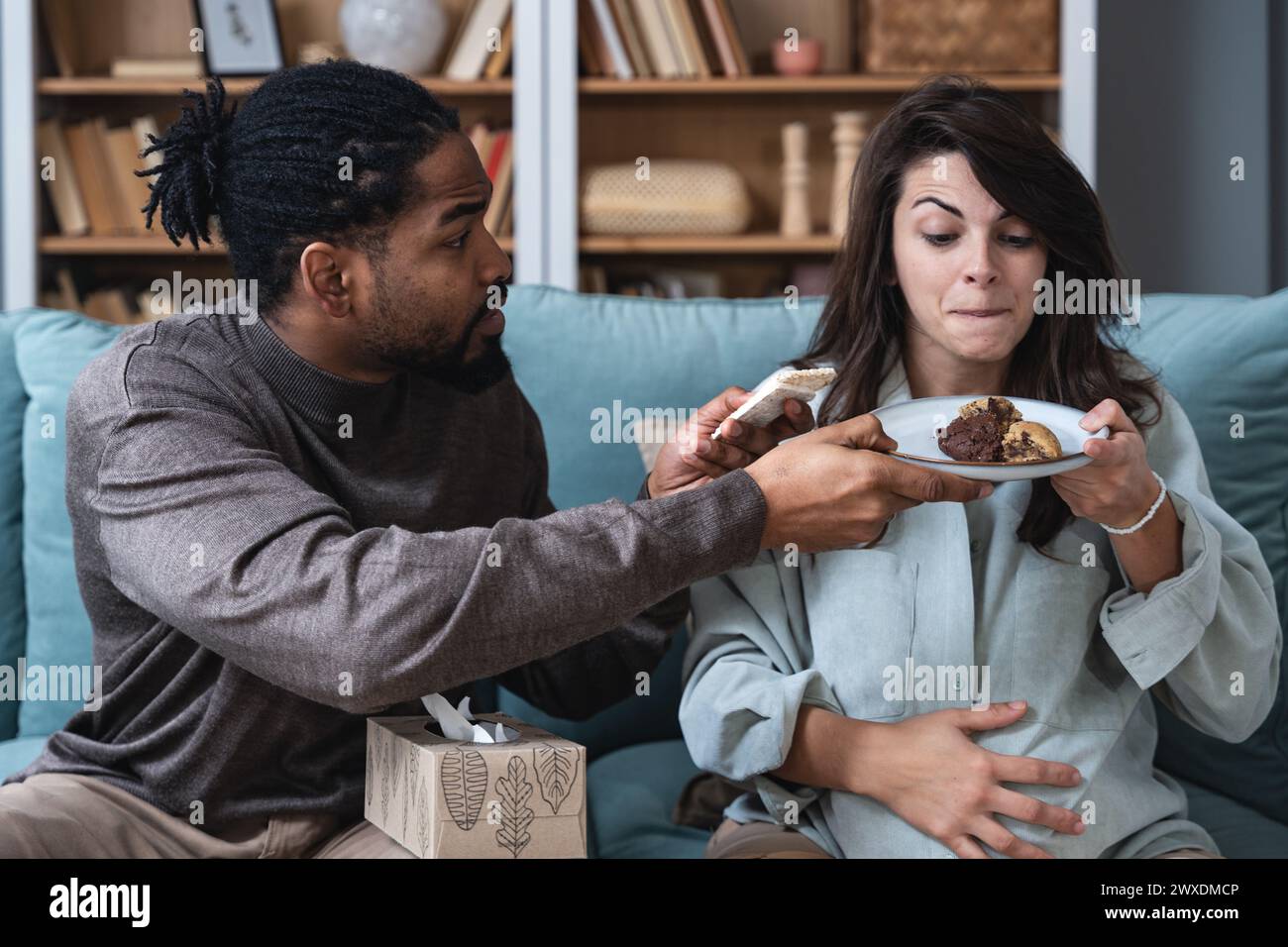 Supporto alla gravidanza e divertimento. Giovane coppia uomo e donna incinta seduta a casa prendere in giro con cibo dolce proibito dal medico. Maschio e femmina supportano AN Foto Stock