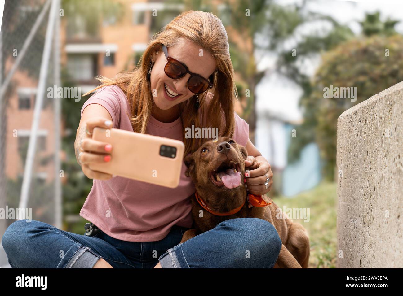 Una donna felice che indossa gli occhiali da sole sorride mentre si fa un selfie con il suo cane adorante. Foto Stock