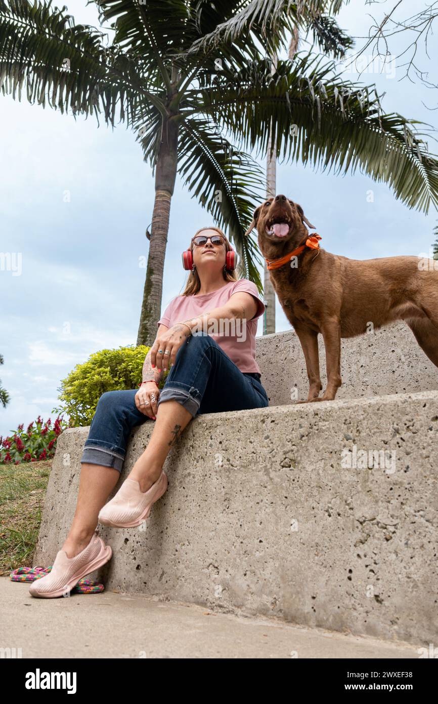 Donna di mezza età seduta all'aperto con cuffie rosse e occhiali da sole, godendosi la giornata di sole con il suo animale domestico. Foto Stock