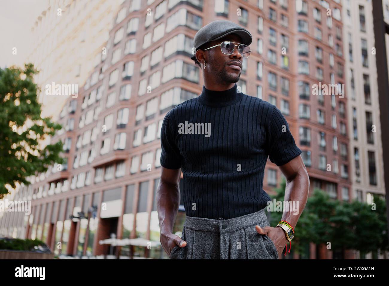 Il bell'elegante uomo afroamericano si erge in strada con un abbigliamento alla moda, indossando un berretto, occhiali da sole e una t-shirt nera, moderna moda maschile Foto Stock