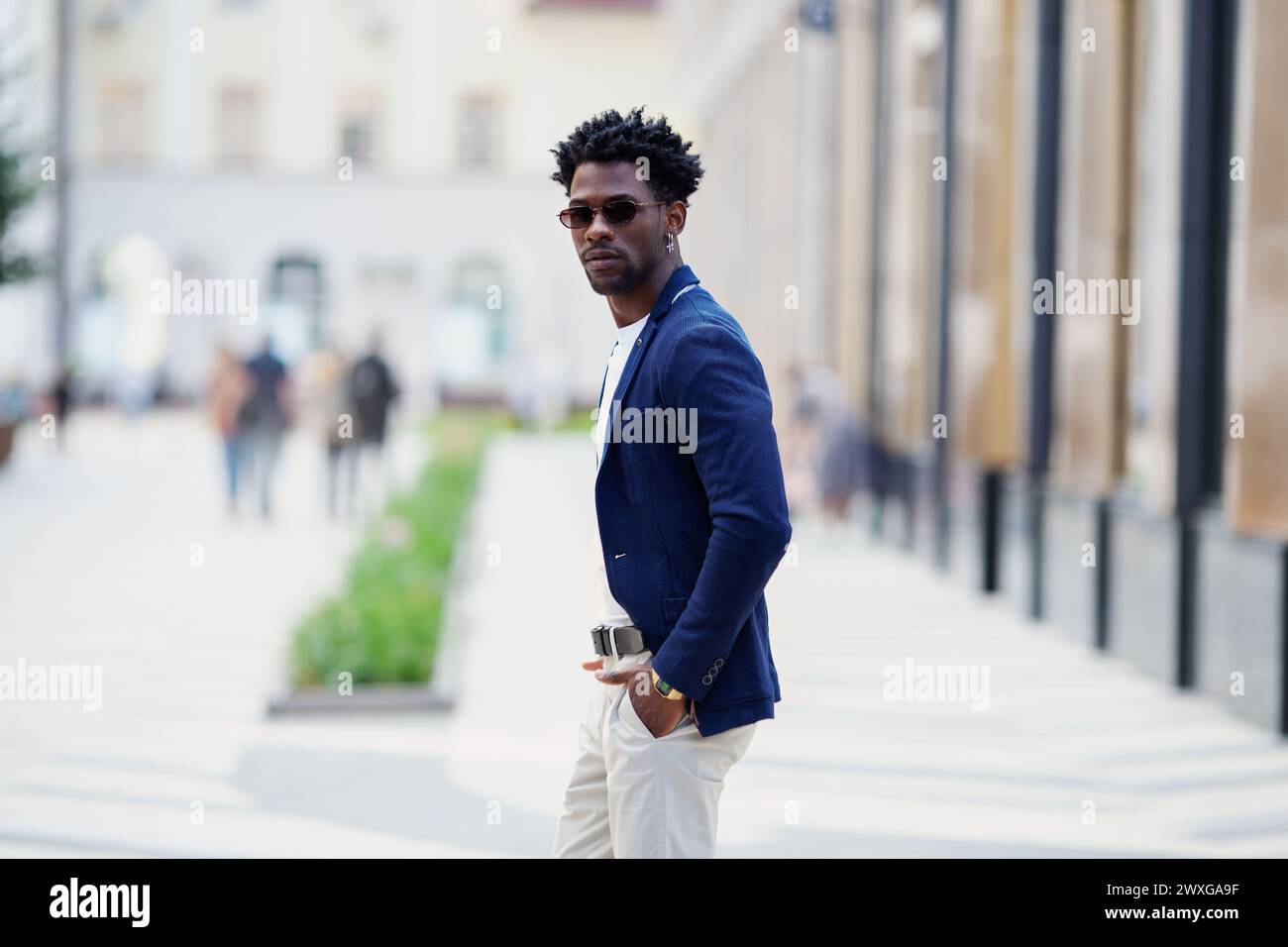 Bell'uomo afroamericano alla moda cammina lungo la strada con eleganti pantaloni bianchi e blazer blu. Tendenza alla moda per lo Street style maschile. Foto Stock