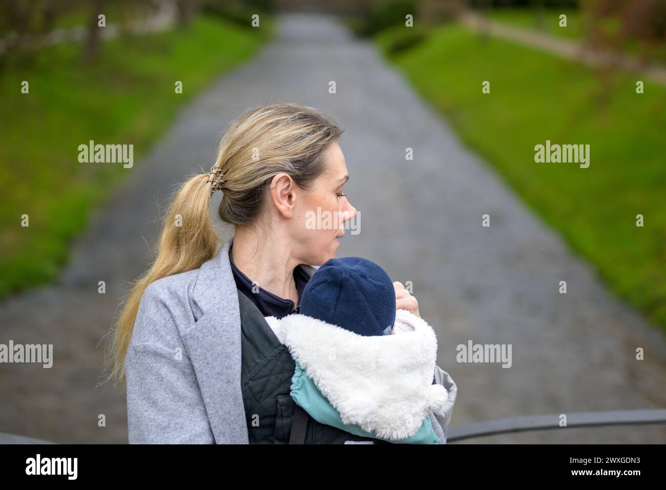 Donna stanca che tiene e trasporta il bambino in un portabicchieri e guarda di fronte a un fiume in un parco Foto Stock