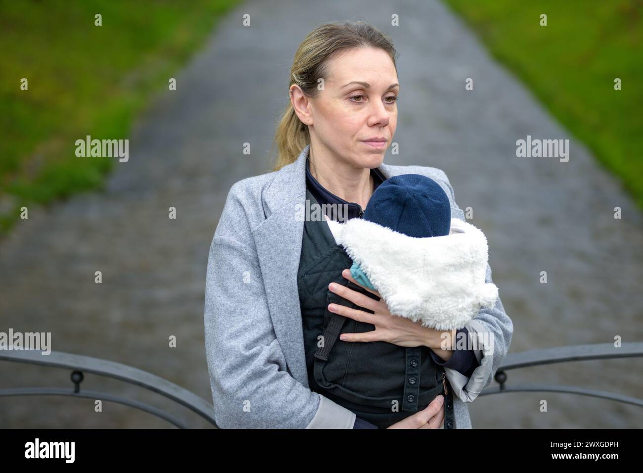 Donna stanca che tiene e trasporta il bambino in un portabicchieri di fronte a un fiume in un parco Foto Stock