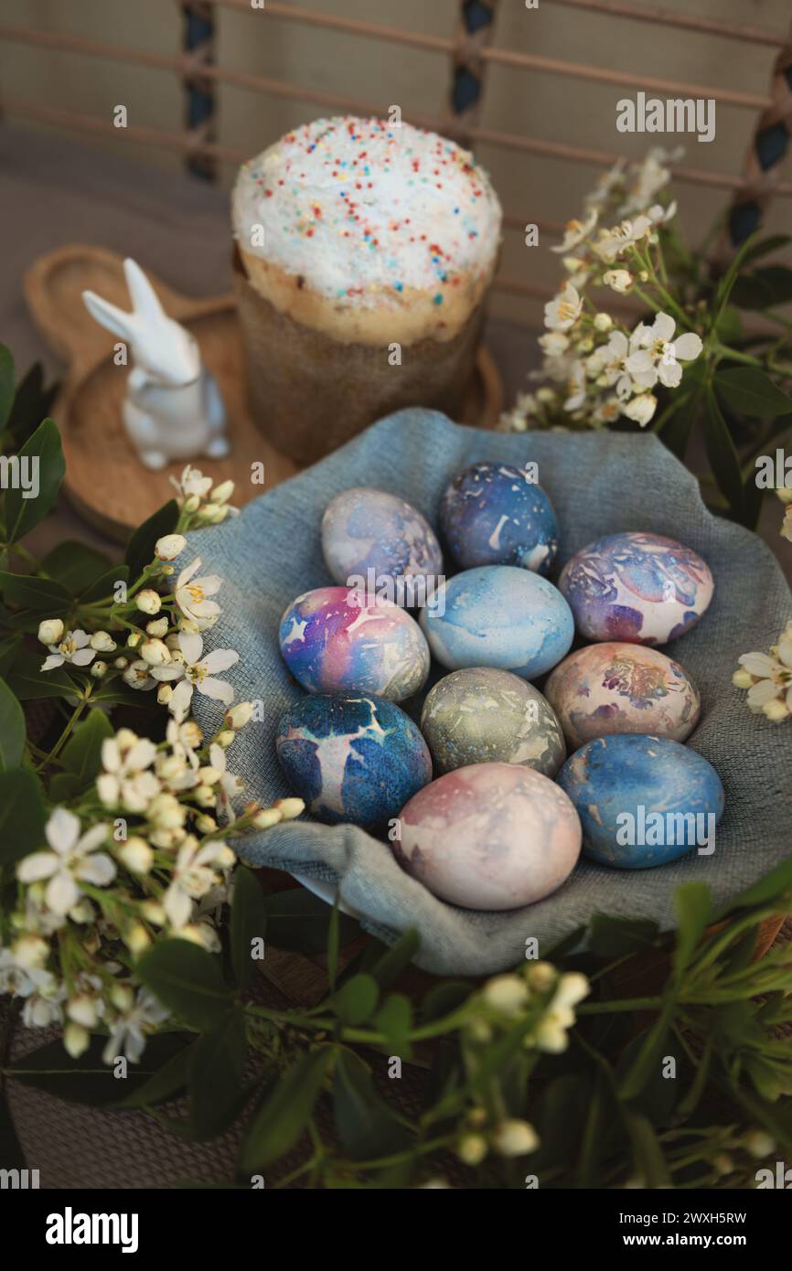 Vista dall'alto delle bellissime uova di Pasqua in marmo con motivi cosmici in una ciotola di tessuto blu circondata da fiori bianchi primaverili. Foto Stock