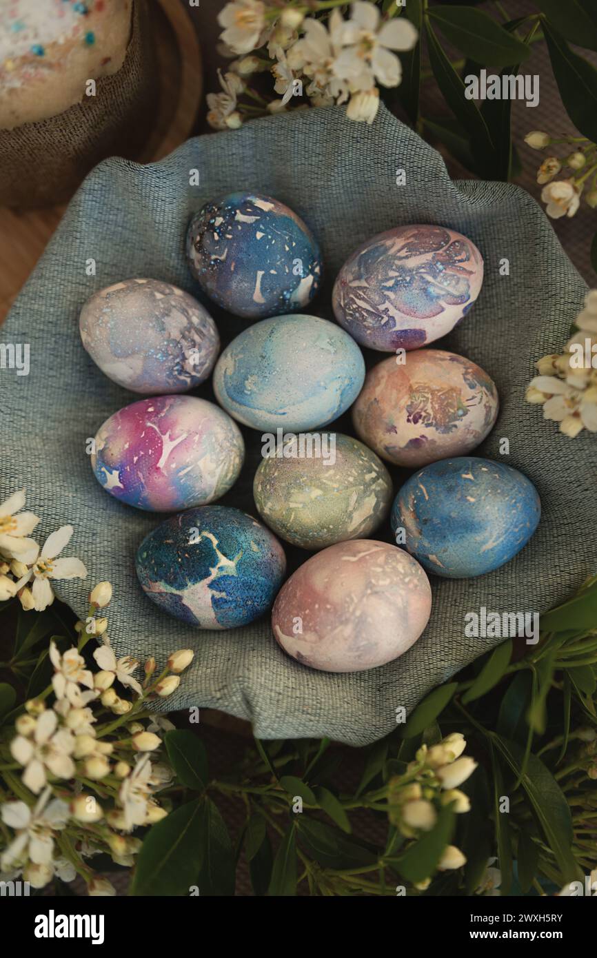 Vista dall'alto delle bellissime uova di Pasqua in marmo con motivi cosmici in una ciotola di tessuto blu circondata da fiori bianchi primaverili. Foto Stock