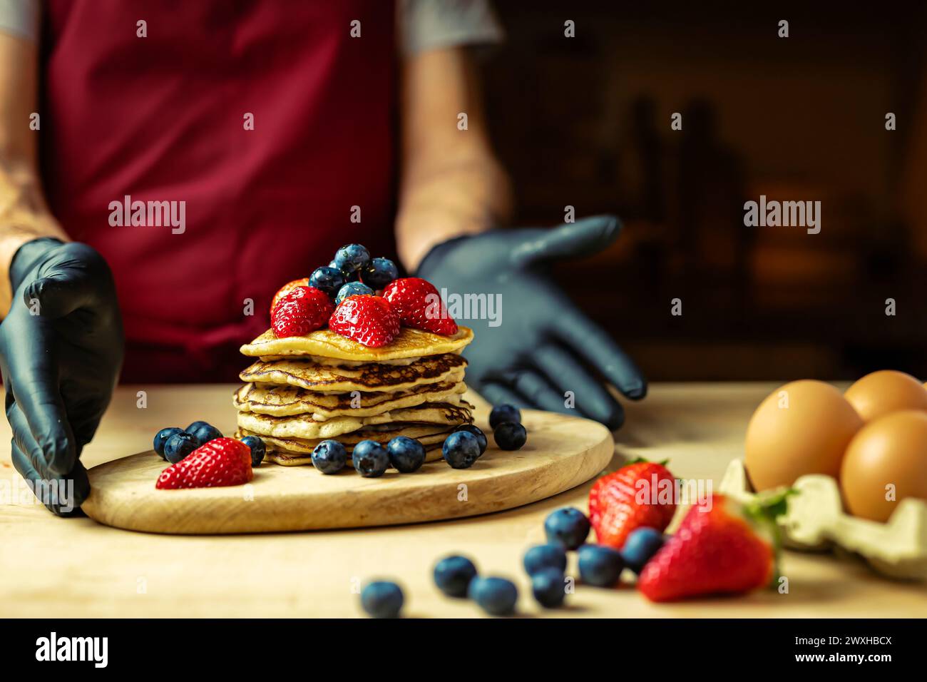 Uomo in grembiule e guanti neri che presenta pancake con panna, fragole e mirtilli su un asse di legno, luce calda Foto Stock