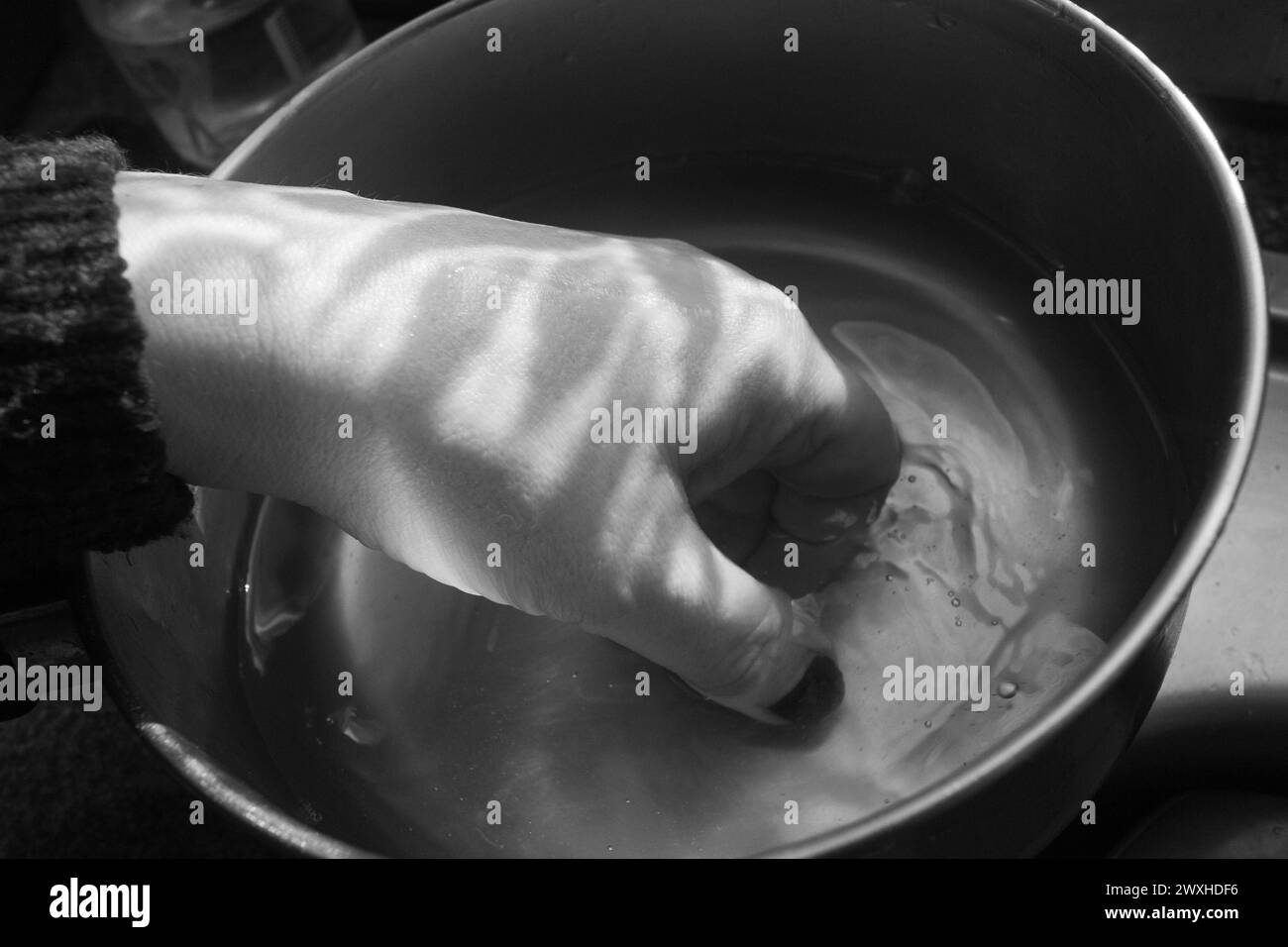 Lavare una mano - foto in bianco e nero di una mano all'interno di una pentola in metallo immersa nell'acqua con luci e ombre contrastanti. Foto Stock