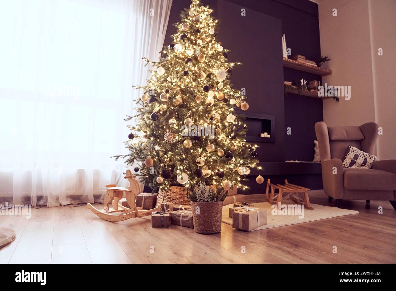 Decorazioni natalizie in un accogliente soggiorno con albero di natale con corde luminose, palle, sedia a dondolo in legno, regali, poltrona, slitta. Sfondo con spazio di copia. Concetto di vigilia di Natale. Foto Stock