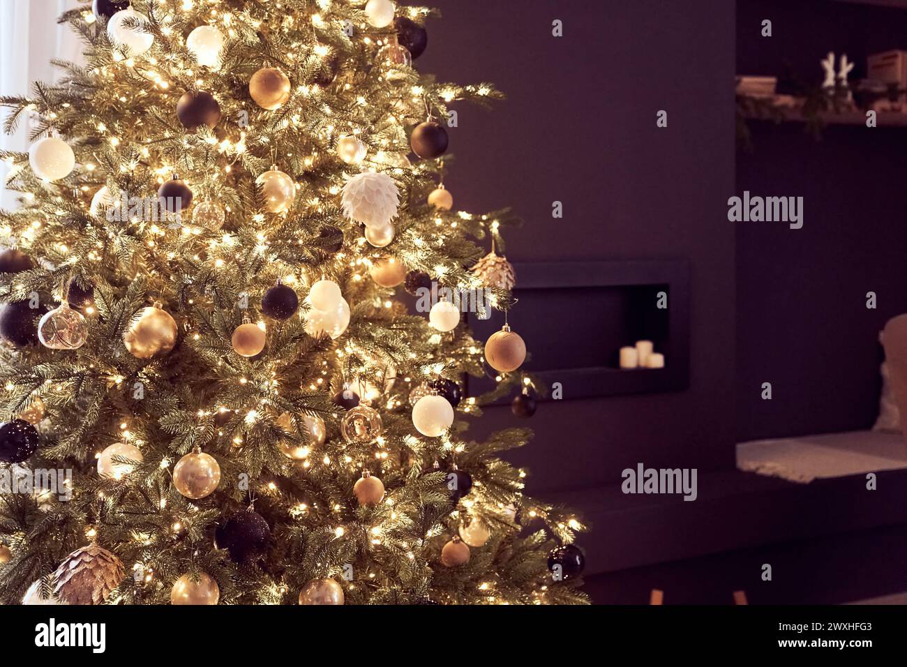 Vista di un albero di Natale con molte decorazioni, luci a corda, bianco, nero, palle d'oro, situato di fronte a un caminetto con candele. Sfondo con spazio di copia. Concetto di Natale, cartolina. Foto Stock