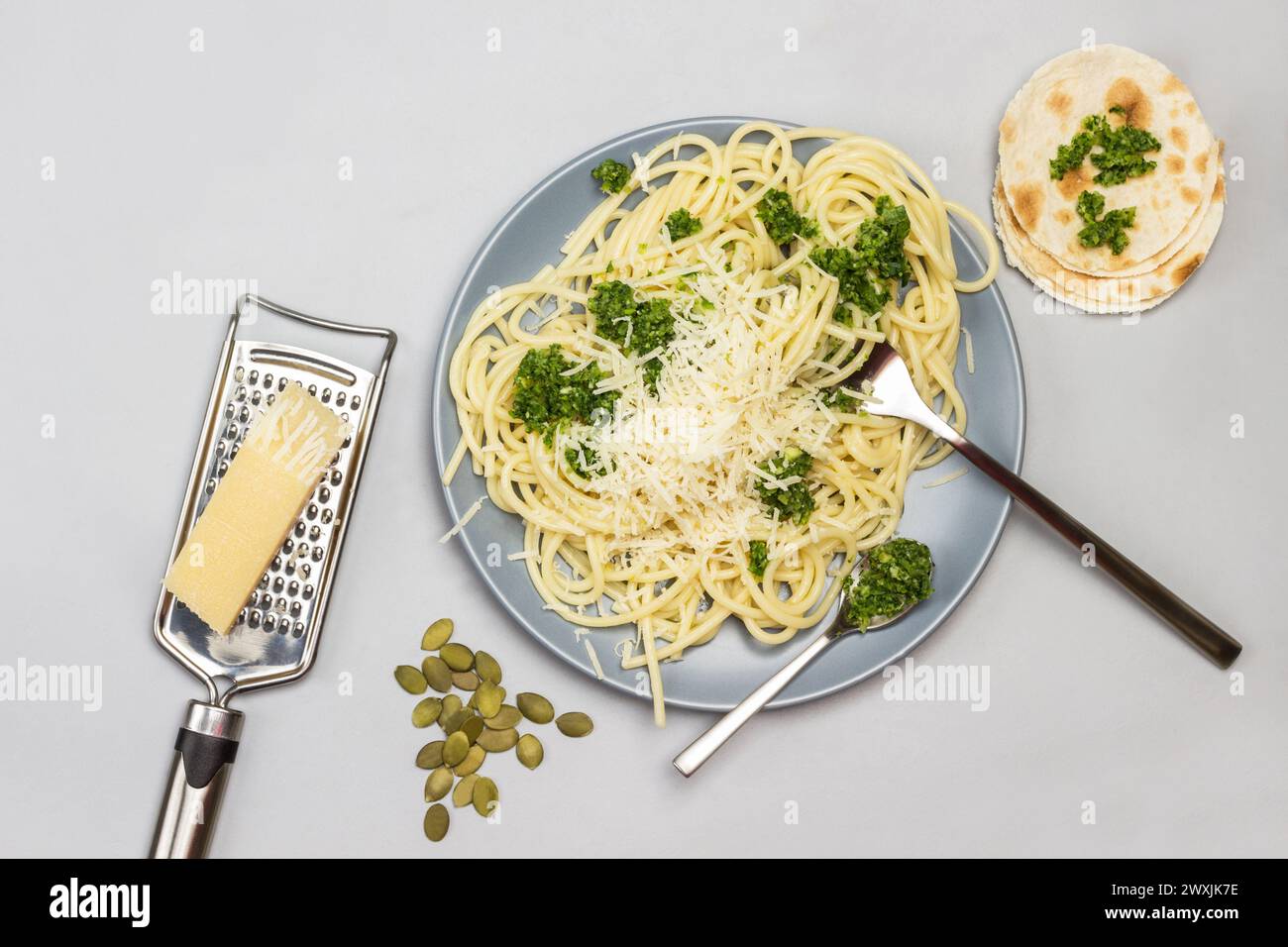 Spaghetti con pasta di broccoli e forchetta e cucchiaio su piatto di ceramica. Parmigiano su grattugia. Focaccine sul tavolo. Sfondo grigio. Posizione piatta Foto Stock