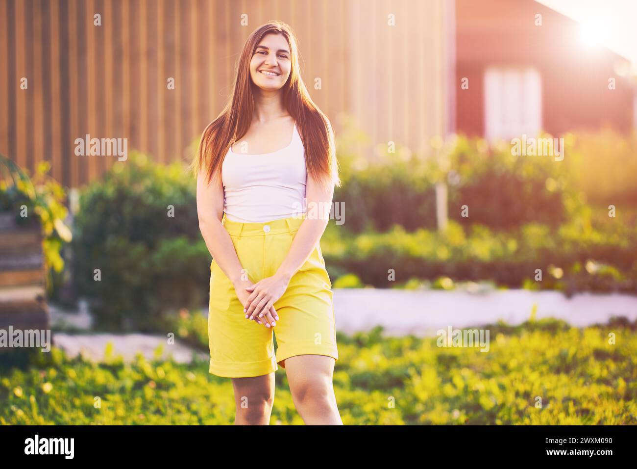 I pantaloncini gialli Bermuda sono indossati da una giovane donna bruna caucasica che posa per fotografare sorridendo mentre si trova nel cortile della casa di campagna alla fine Foto Stock
