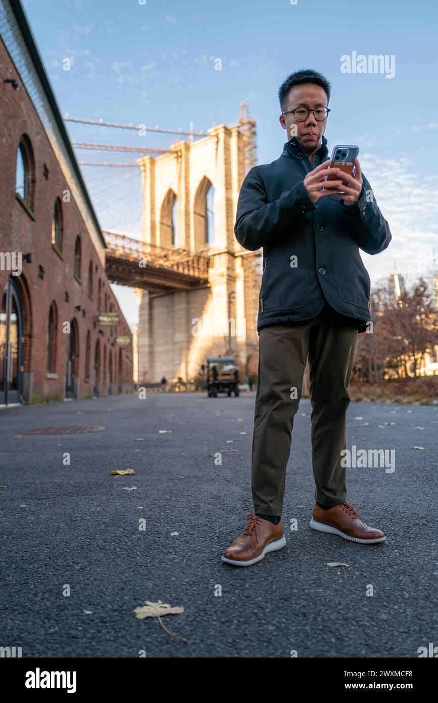 Uomo concentrato che usa lo smartphone accanto all'iconico ponte di Brooklyn Foto Stock