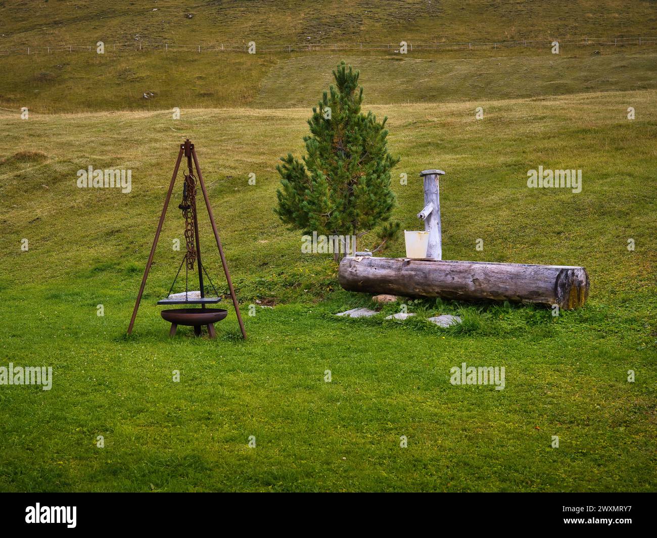 Tradizionale treppiede da cucina all'aperto sopra un piccolo camino accanto a una vasca d'acqua in legno su verdi pascoli di montagna. Rustica scena rurale con una semplice rocca Foto Stock