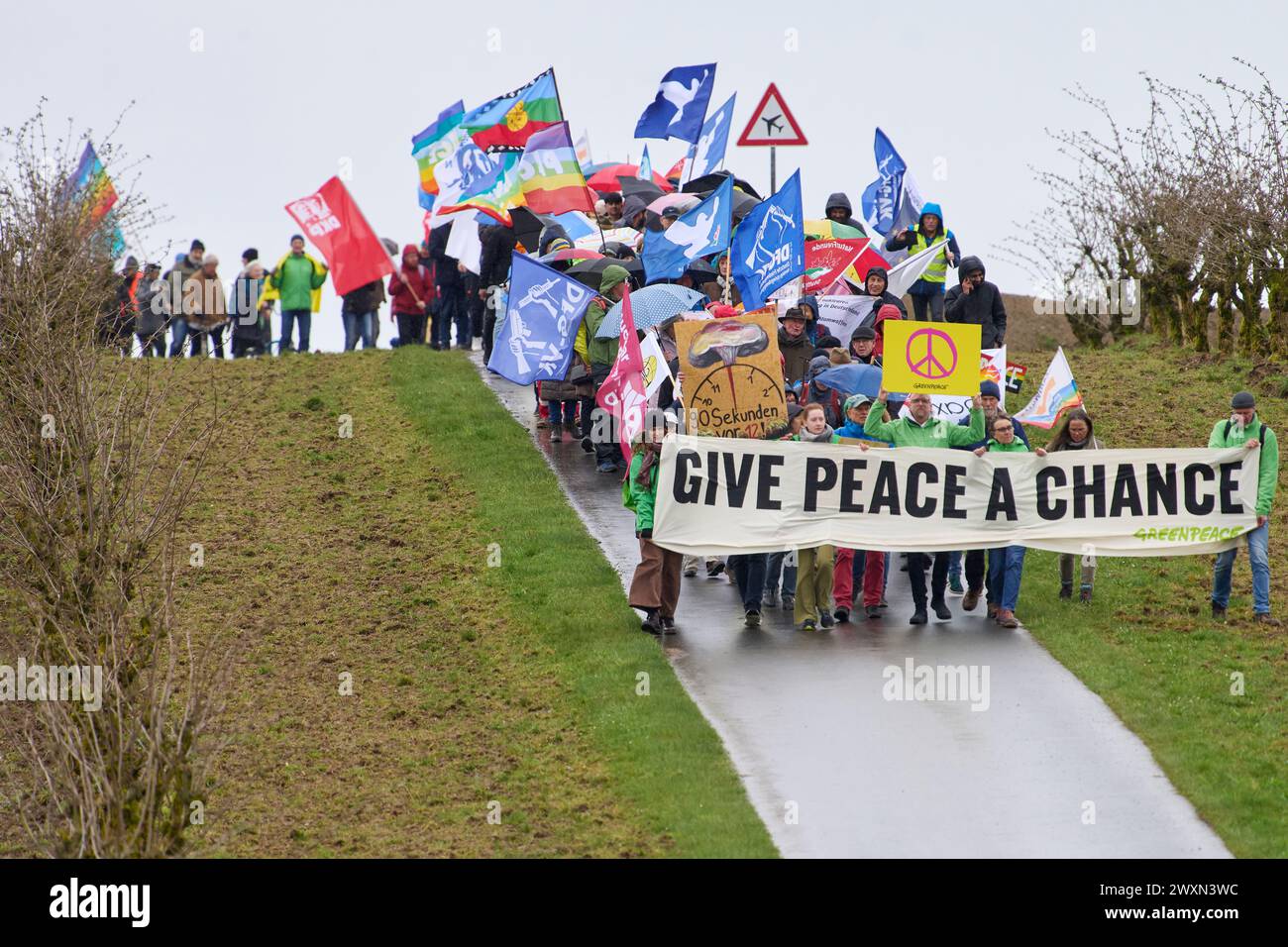 01 aprile 2024, Renania-Palatinato, Büchel: I partecipanti alla marcia di Pasqua presso la base aerea di Büchel (distretto di Cochem-Zell) camminano lungo la recinzione esterna della base, dove si dice che siano immagazzinate armi nucleari statunitensi. Lo striscione recita: "Date Una possibilità alla pace”. Foto: Thomas Frey/dpa Foto Stock
