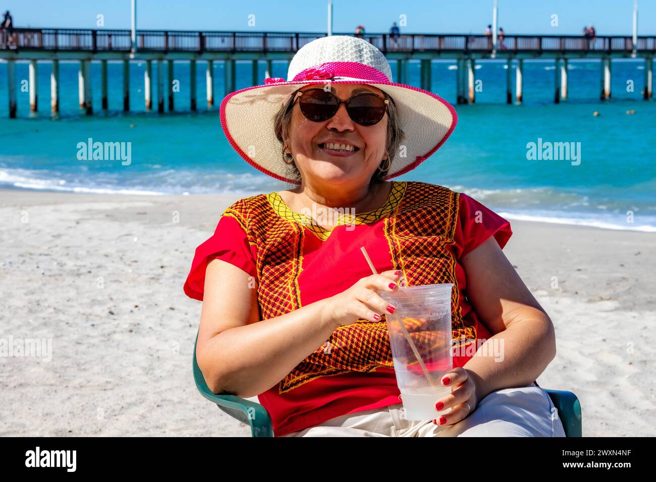 Donna messicana sorridente seduta rinfrescandosi bevendo acqua di cocco in un bicchiere, un molo e l'oceano sullo sfondo, cappello, occhiali da sole e camicetta rossa, godendosi su Foto Stock Donna messicana sorridente seduta rinfrescandosi bevendo acqua di cocco in un bicchiere, un molo e l'oceano sullo sfondo, cappello, occhiali da sole e camicetta rossa, godendosi su Foto Stock