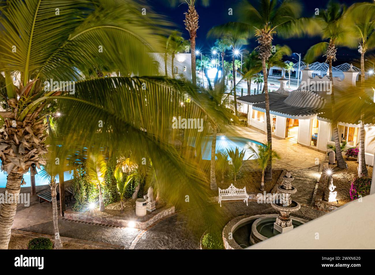 Palme sulla terrazza con vista notturna, area relax all'aperto presso il resort con piscina, fontana della cava, edificio e strada con luci accese, vista dalla ba Foto Stock