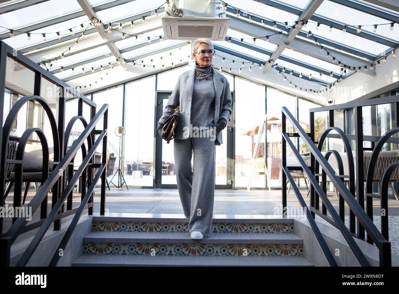 Donna matura attraente ed elegante in pantalone grigio con giacca e dolcevita, accessoriata con borsa e scarpe bianche. Il look perfetto per succ Foto Stock