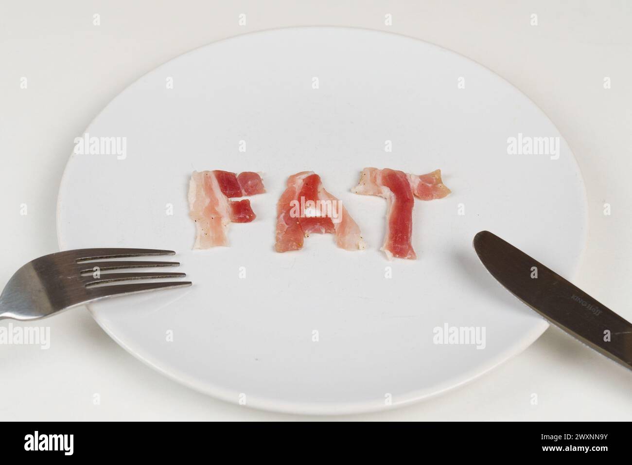 Un piatto con sopra la parola grasso, accompagnato da una forchetta e un coltello, che simboleggiano abitudini alimentari malsane. Foto Stock