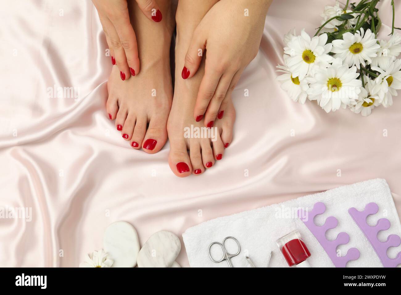 Donna con eleganti unghie rosse dopo la pedicure, attrezzi e fiori di camomilla su tessuto di seta chiaro, vista dall'alto Foto Stock