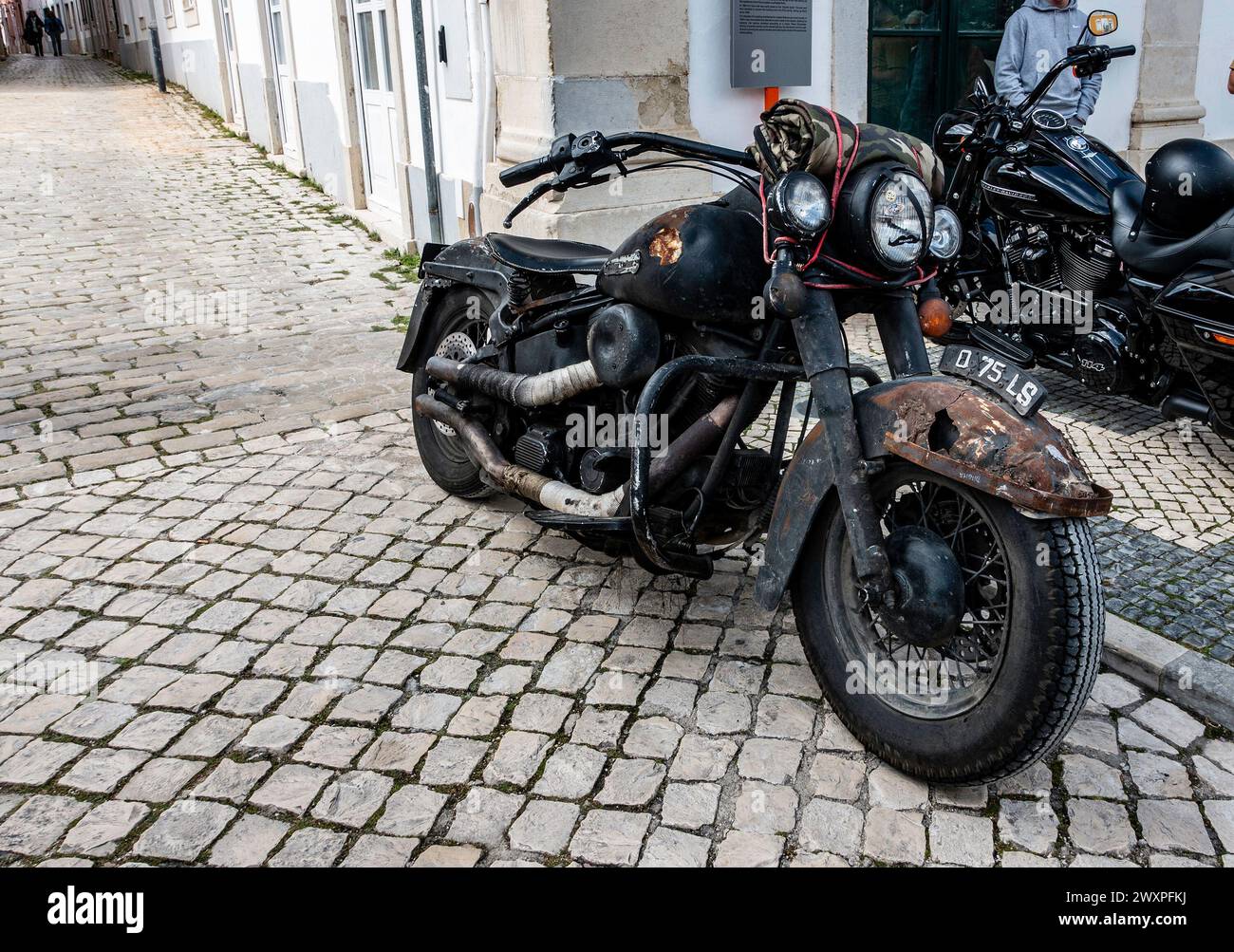 Una motocicletta arrugginita d'epoca con una patina distinta si trova parcheggiata su una vecchia strada acciottolata, a Loule, Portogallo. Foto Stock