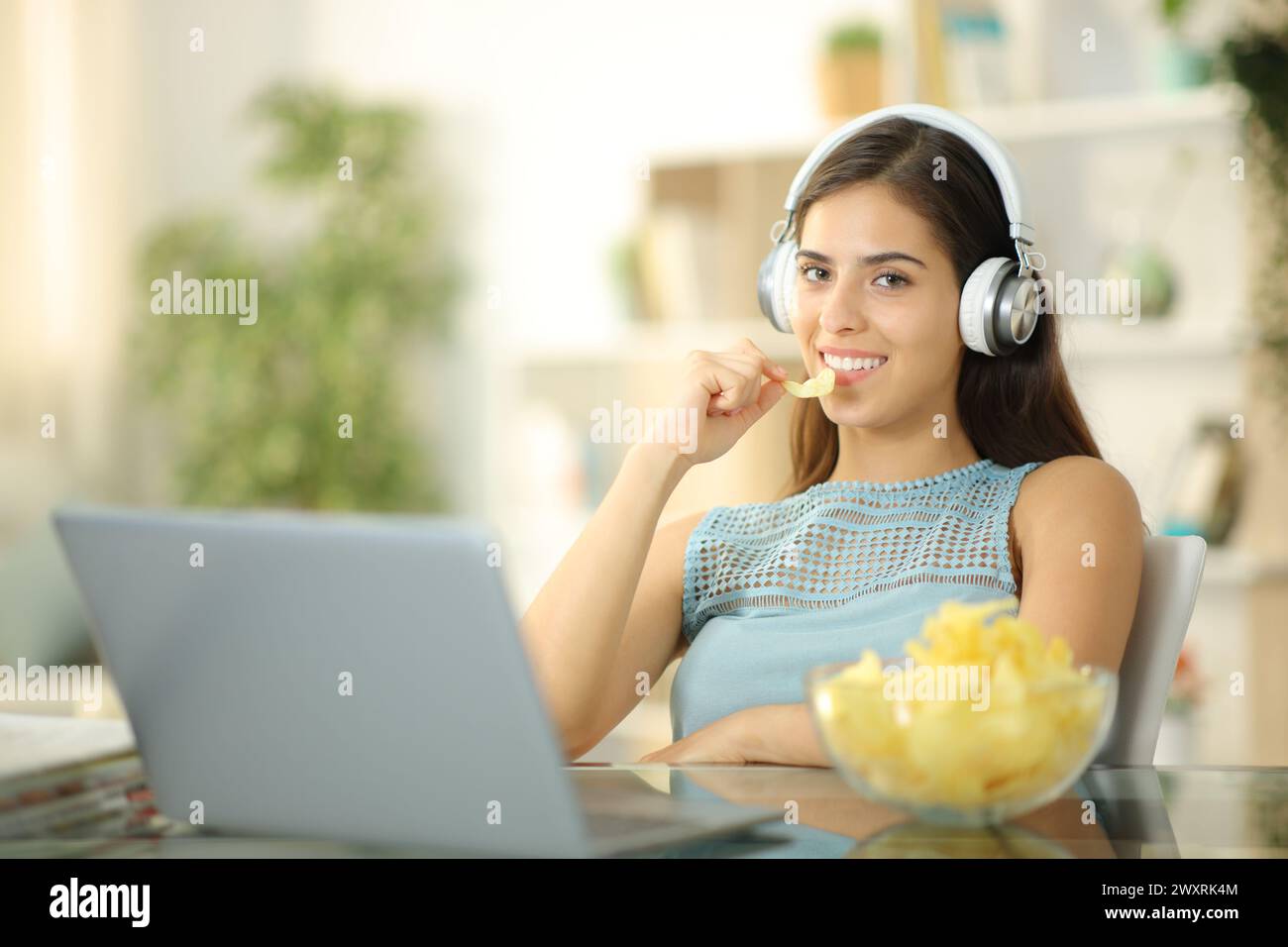 Donna con laptop e cuffie che mangia patatine che guarda la macchina fotografica a casa Foto Stock