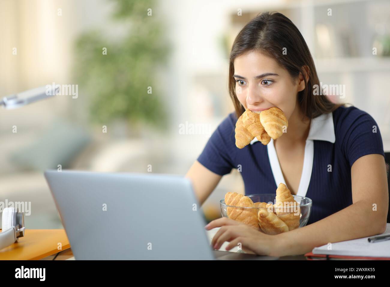 E-learning studentesco goloso con un portatile che mangia molti croissant a casa Foto Stock