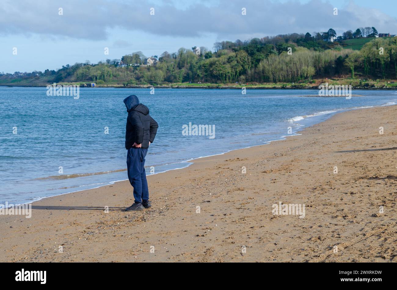 Crawfordsburn County Down Northern Ireland marzo 30 2024 - uomo incappucciato sulla spiaggia che guarda al mare contemplando la sua vita Foto Stock