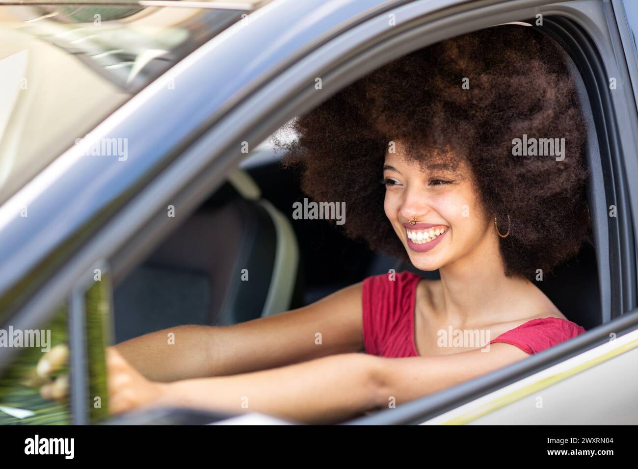 Sorridente eccitata giovane donna etnica con auto di guida dai capelli afro scuri il giorno d'estate in città Foto Stock