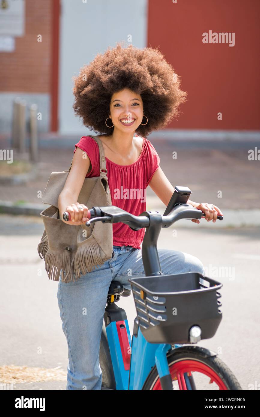 Donna afroamericana positiva in abiti casual con acconciatura afro e borsa sorridente e guardando la macchina fotografica mentre si guida in bici elettrica su City st Foto Stock