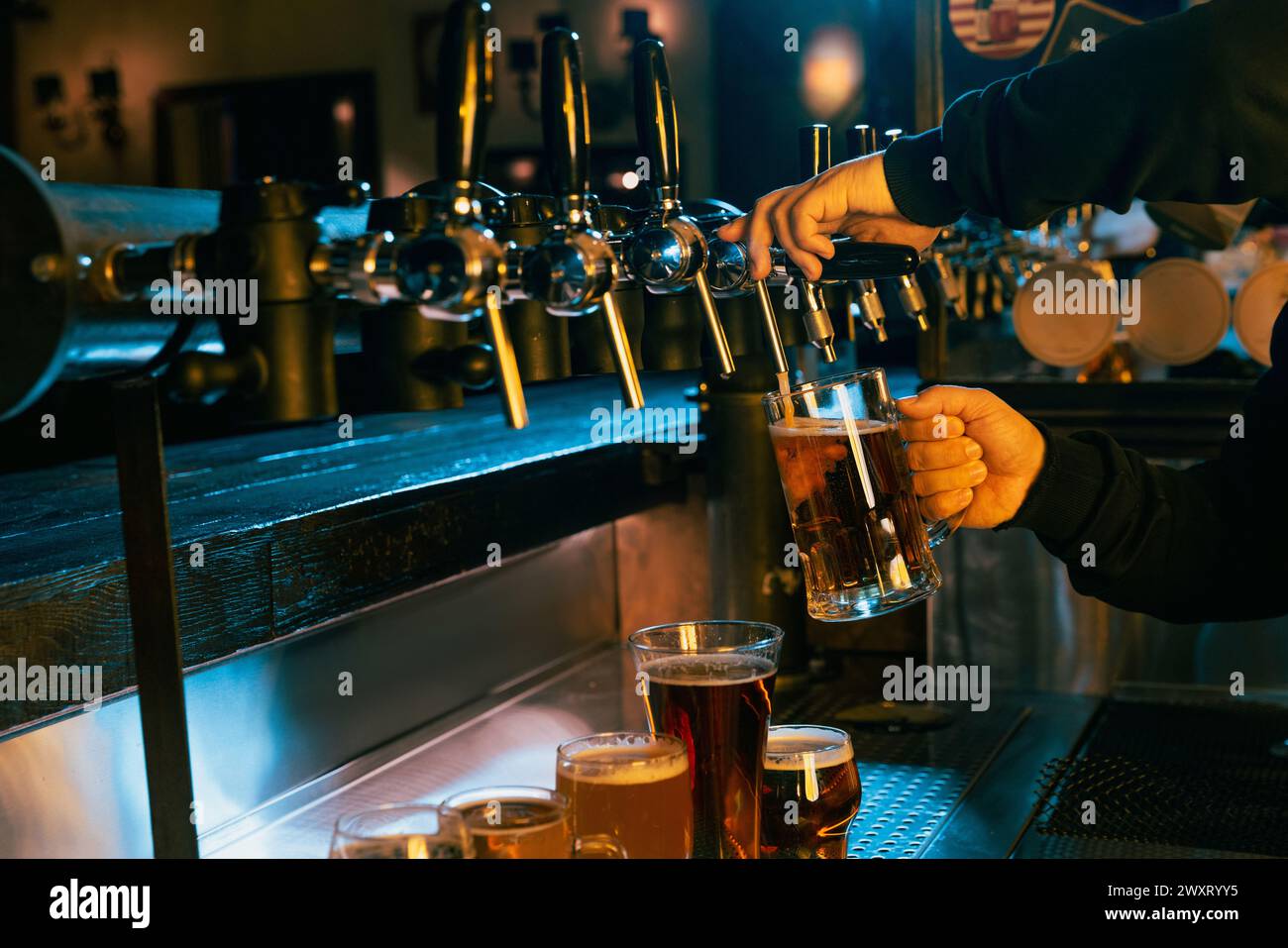 Estrazione a mano di birra lager fresca e rinfrescante dalla spina con varie altre birre esposte al bar ben illuminato. Serve birra artigianale alla spina. Foto Stock