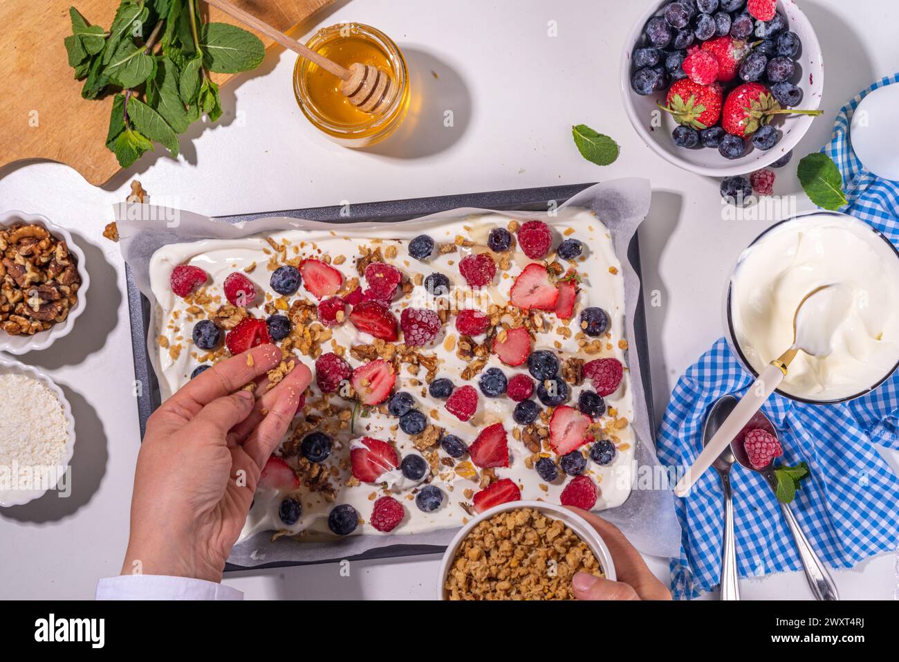 Preparazione della cottura con sfondo di corteccia di yogurt congelato con yogurt greco, bacche fresche, muesli, miele, salsa al cioccolato, con vassoio da forno deco. Vista dall'alto della donna Foto Stock