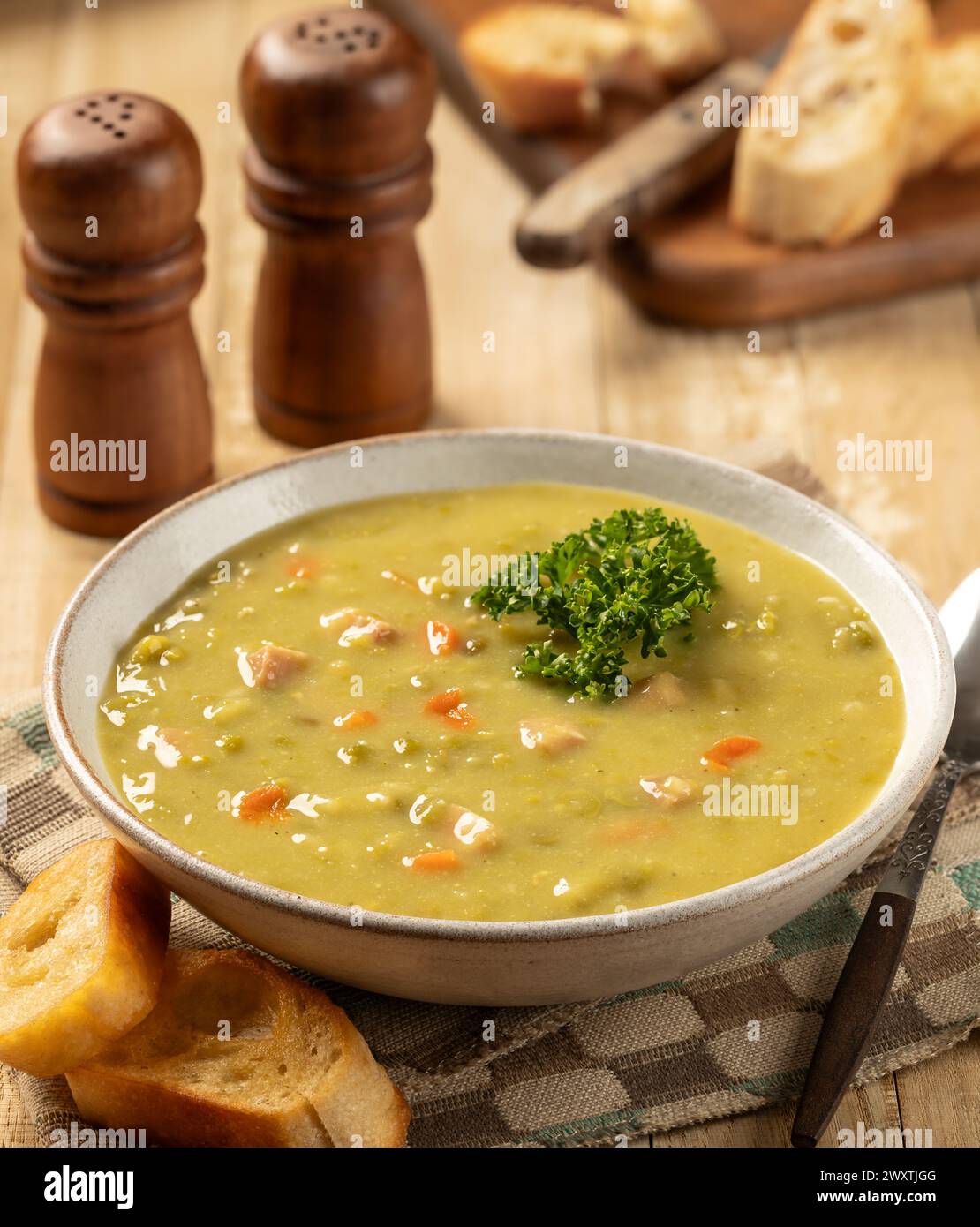 Ciotola di zuppa di piselli con prosciutto e carote guarnite con prezzemolo e baguette tostate su un tavolo di legno Foto Stock