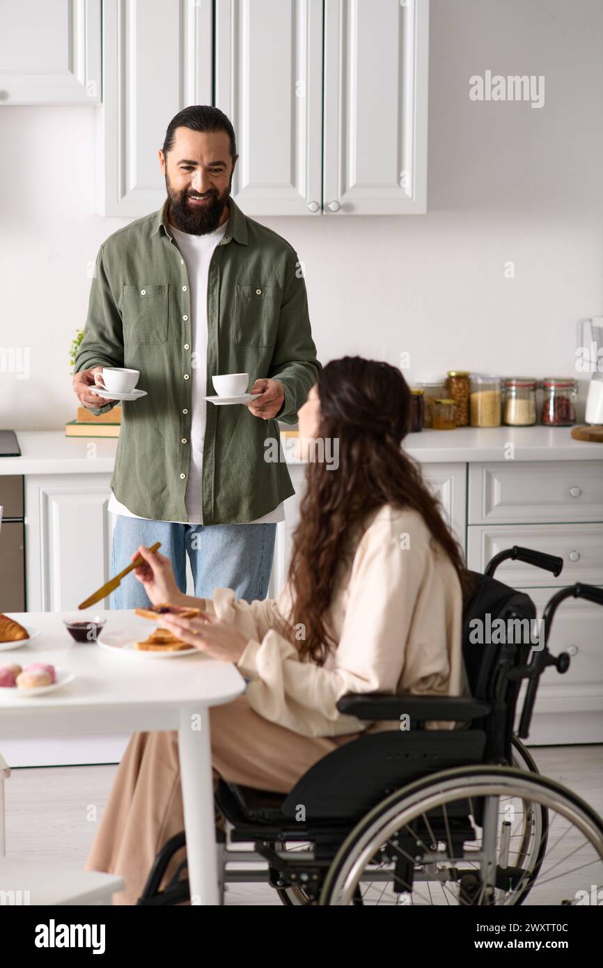 bella donna allegra con disabilità in sedia a rotelle che fa colazione con il marito barbuto Foto Stock