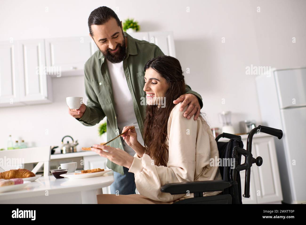 bella donna allegra con disabilità in sedia a rotelle che fa colazione con il marito barbuto Foto Stock