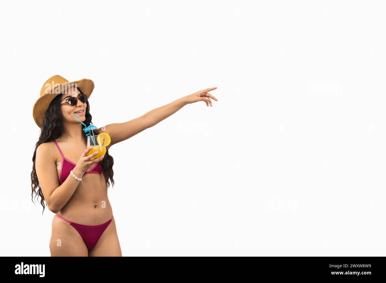 Copyspace giovane donna latina in bikini si gode un cocktail sulla spiaggia mentre punta verso l'alto e verso destra con la mano, forse indicando Foto Stock