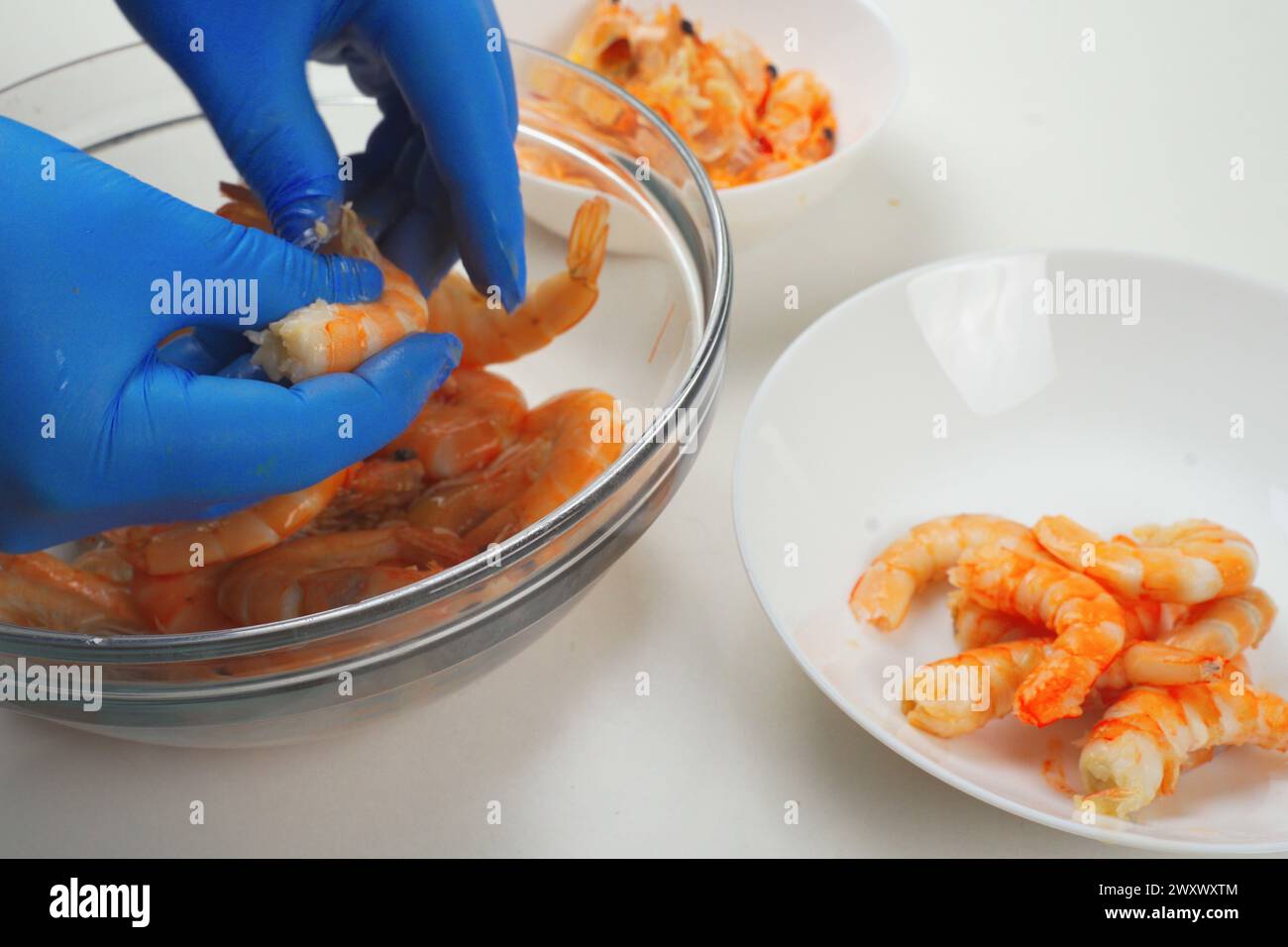 Una persona che indossa guanti blu sta sbucciando i gamberi su una ciotola di vetro trasparente accanto a un piatto bianco con gamberi sbucciati, suggerendo di preparare il pasto in una cupola Foto Stock