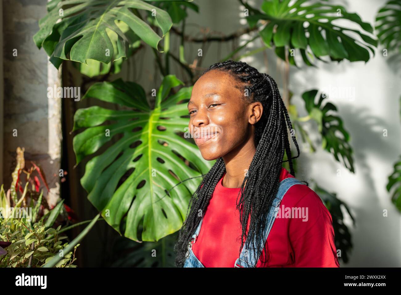 Una giovane donna afroamericana felice sta accanto alla finestra, crogiolandosi alla luce del sole con un sorriso sul viso, e si sente gioia. Foto Stock