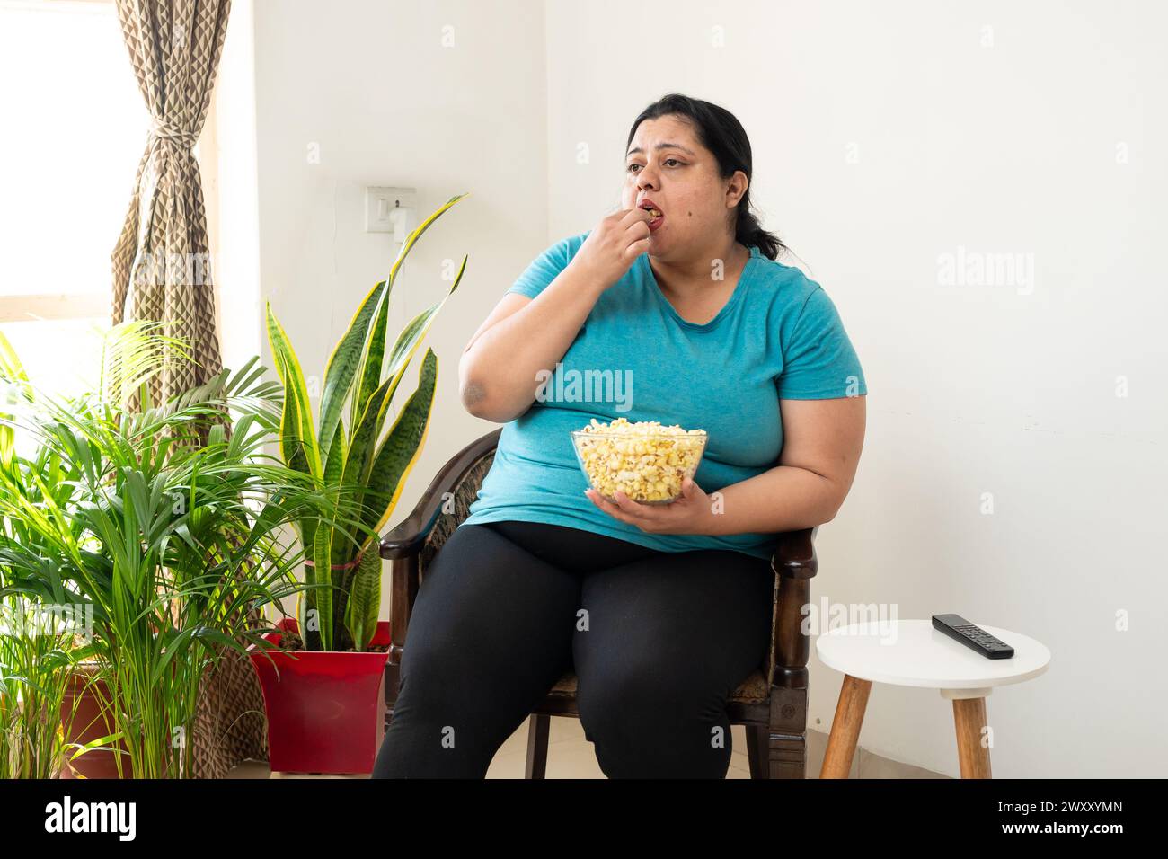 Donna indiana grassa in sovrappeso seduta sulla sedia a mangiare popcorn a casa. Le donne più grandi possono gustare spuntini. Concetto di alimentazione malsana. Foto Stock