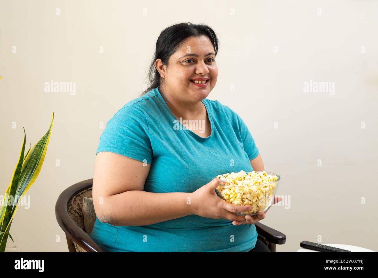Donna indiana grassa in sovrappeso seduta su una sedia con una ciotola di popcorn in mano. Inoltre, una donna di taglia superiore gusta spuntini. Concetto di alimentazione malsana. Foto Stock
