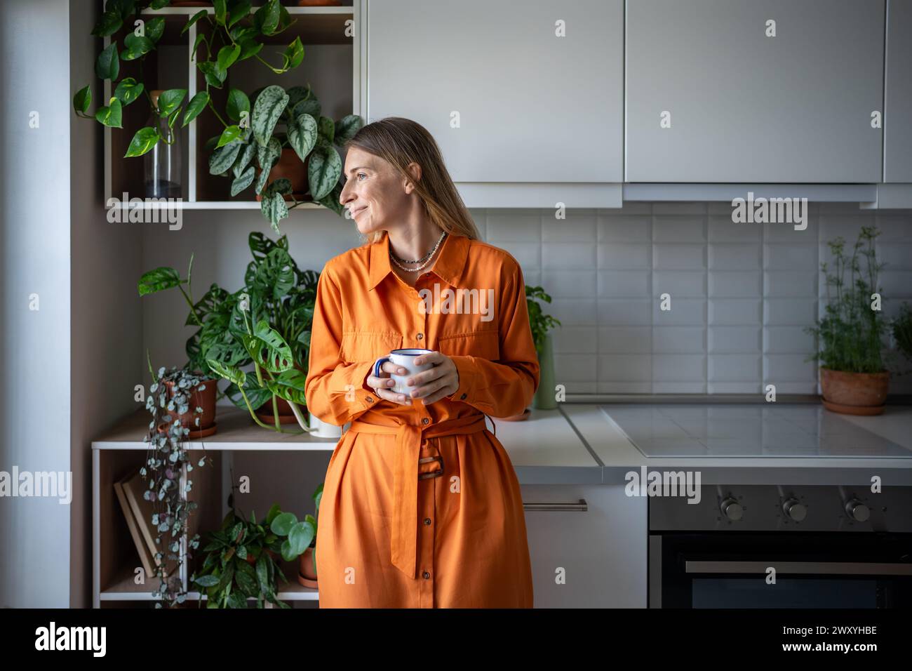 Donna sorridente e spensierata che beve tè in cucina tra le piante d'appartamento, guardando fuori dalla finestra. Vita lenta. Foto Stock