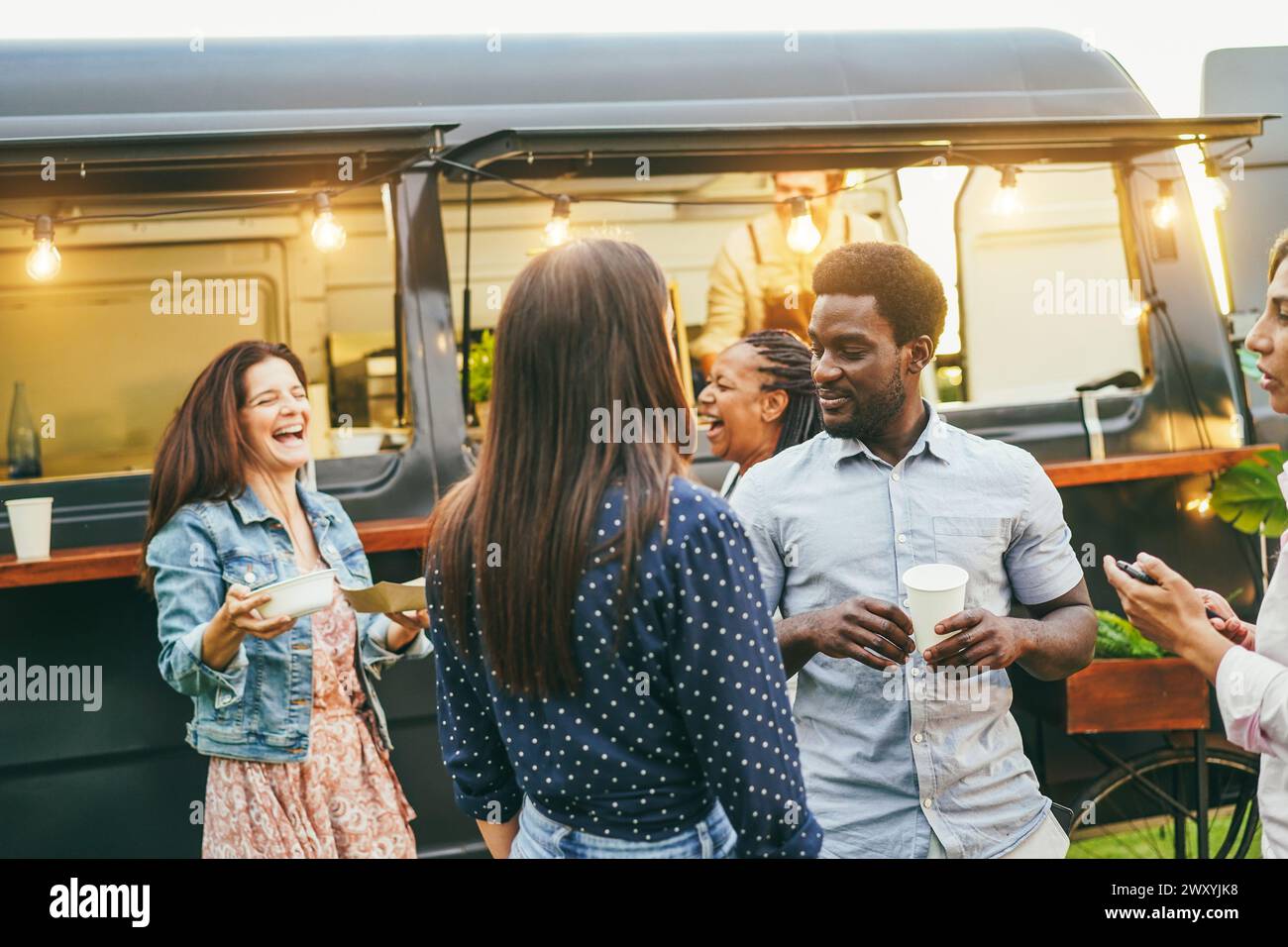 Persone multirazziali che mangiano e bevono di fronte al food truck all'aperto - amici multigenerazionali che cenano fuori durante l'estate Foto Stock