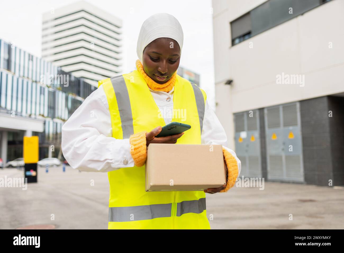 Una donna musulmana africana sorridente che indossa un hijab e un giubbotto di sicurezza contiene un pacchetto e utilizza il suo smartphone in un ambiente urbano. Foto Stock